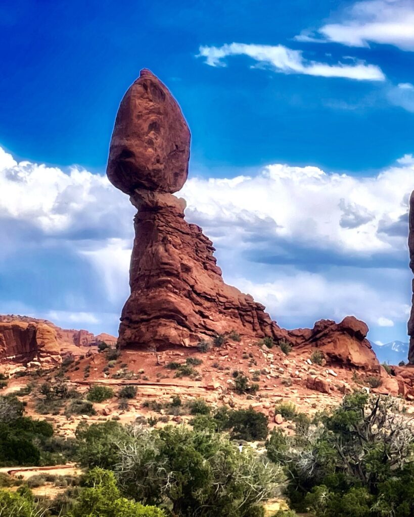 Balanced Rock in Arches National Park, een reusachtige ovale steen die op een smalle rotspilaar balanceert onder een blauwe hemel met wolken.
